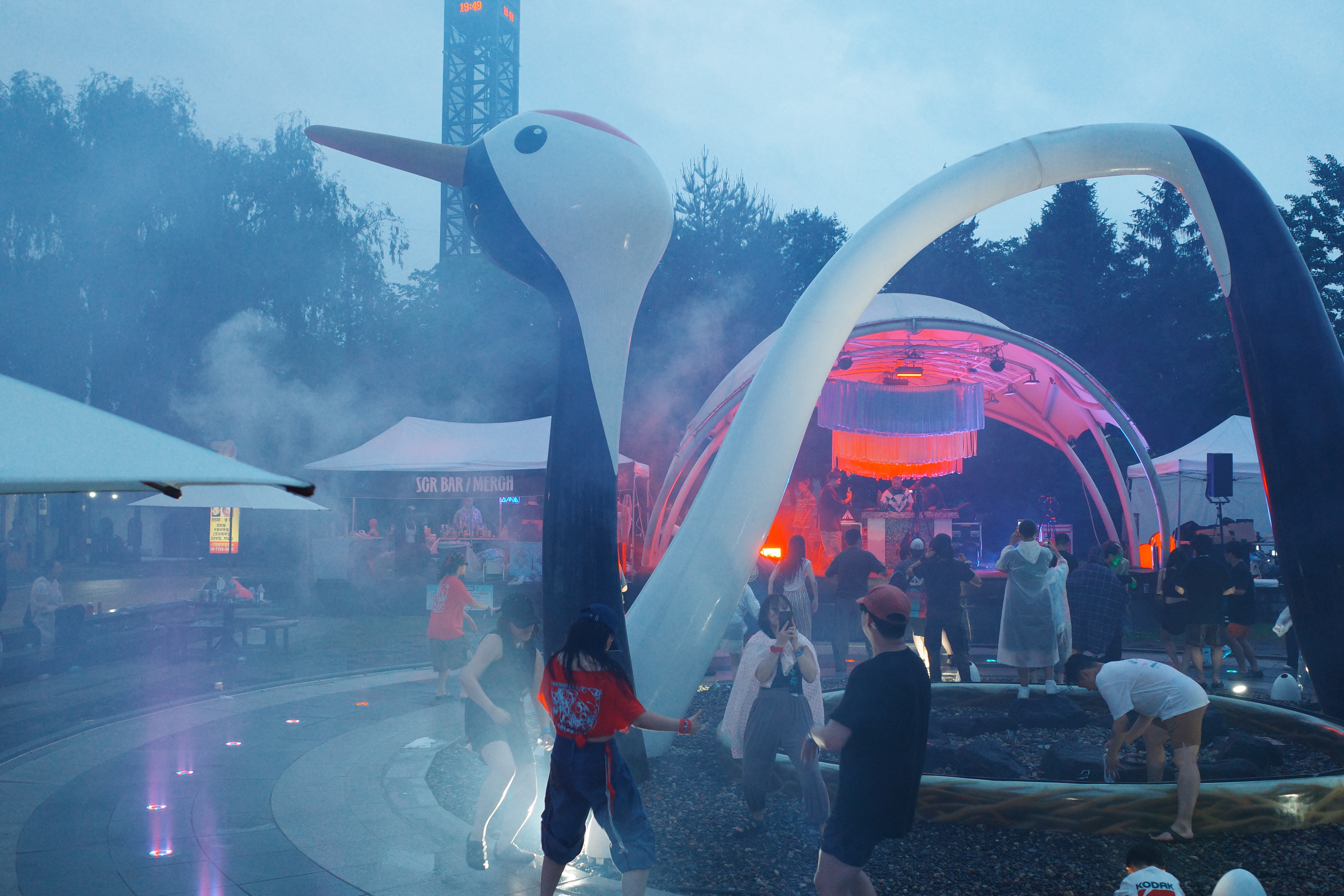 People gathered at an outdoor evening festival under misty rain, near a large bird-shaped sculpture arch